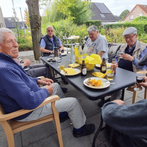 Mehrere ältere Männer sitzen gemeinsam an einem Tisch auf einer Terrasse vor einem Wohngebäude. Auf dem Tisch stehen Getränke, Gläser und verschiedene Saucen