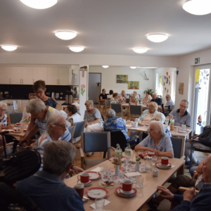 Zahlreiche ältere Menschen sitzen gemeinsam an mehreren Tischen in einem hellen Gemeinschaftsraum. Auf den Tischen stehen Kaffeetassen, Teller, Gläser und Getränke.
