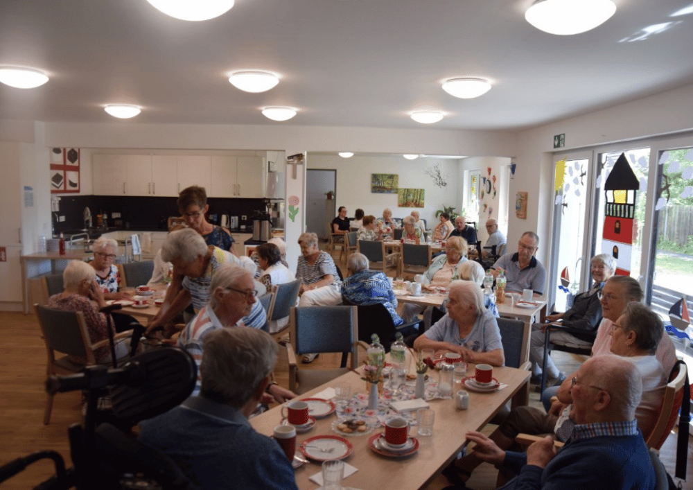 Zahlreiche ältere Menschen sitzen gemeinsam an mehreren Tischen in einem hellen Gemeinschaftsraum. Auf den Tischen stehen Kaffeetassen, Teller, Gläser und Getränke.