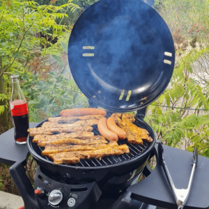 Auf einem geöffneten runden Gasgrill liegen mehrere Stücke Fleisch und Würstchen. Rauch steigt vom Grill auf, daneben stehen eine Getränkeflasche und eine Grillzange