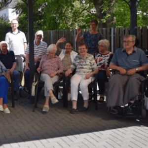 Eine größere Gruppe älterer Menschen sitzt und steht gemeinsam unter einem überdachten Pavillon im Garten. Mehrere Personen lächeln in die Kamera und heben ihre Hände.