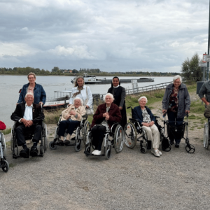 Eine Gruppe älterer Menschen und Begleitpersonen steht und sitzt mit Rollstühlen und Rollatoren an einem Flussufer. Im Hintergrund fährt ein Frachtschiff auf dem Wasser unter bewölktem Himmel.