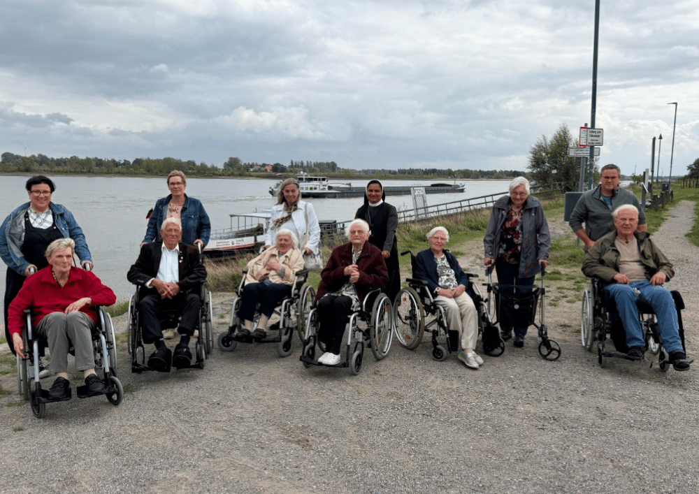 Eine Gruppe älterer Menschen und Begleitpersonen steht und sitzt mit Rollstühlen und Rollatoren an einem Flussufer. Im Hintergrund fährt ein Frachtschiff auf dem Wasser unter bewölktem Himmel.