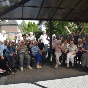 Eine größere Gruppe älterer Menschen sitzt und steht gemeinsam unter einem überdachten Pavillon im Garten. Mehrere Personen lächeln in die Kamera und heben ihre Hände.