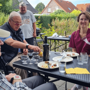 Fünf Männer sitzen an einem Tisch auf einer Terrasse und essen und trinken gemeinsam. Teller, Flaschen, Gläser und gelbe Servietten stehen auf dem Tisch, im Hintergrund ist ein Garten zu sehen