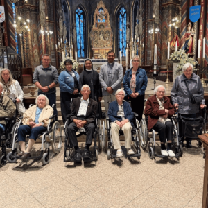 Eine Gruppe älterer Menschen und Begleitpersonen posiert im reich verzierten Innenraum einer Kirche vor dem Altar. Mehrere Personen sitzen im Rollstuhl, weitere stehen dahinter.