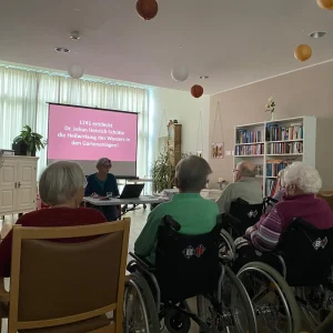 Bewohnerinnen und Bewohner verfolgen einen Vortrag zur Geschichte des Forstgartens in Kleve. Auf der Leinwand wird die Entdeckung der Heilwirkung des Wassers im Jahr 1741 thematisiert