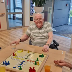 Vier ältere Menschen sitzen um ein großes Mensch-ärgere-dich-nicht-Spielbrett. Eine Frau und ein Mann lächeln in die Kamera.