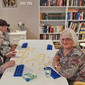Zwei ältere Frauen und eine jüngere Person sitzen an einem Tisch mit ausgelegten Zahlenplättchen und blauen Spielsteinständern.