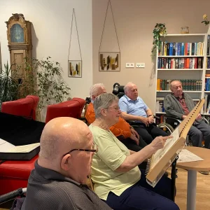 Eine ältere Frau spielt ein Saiteninstrument aus Holz. Mehrere ältere Menschen sitzen neben ihr und hören zu.