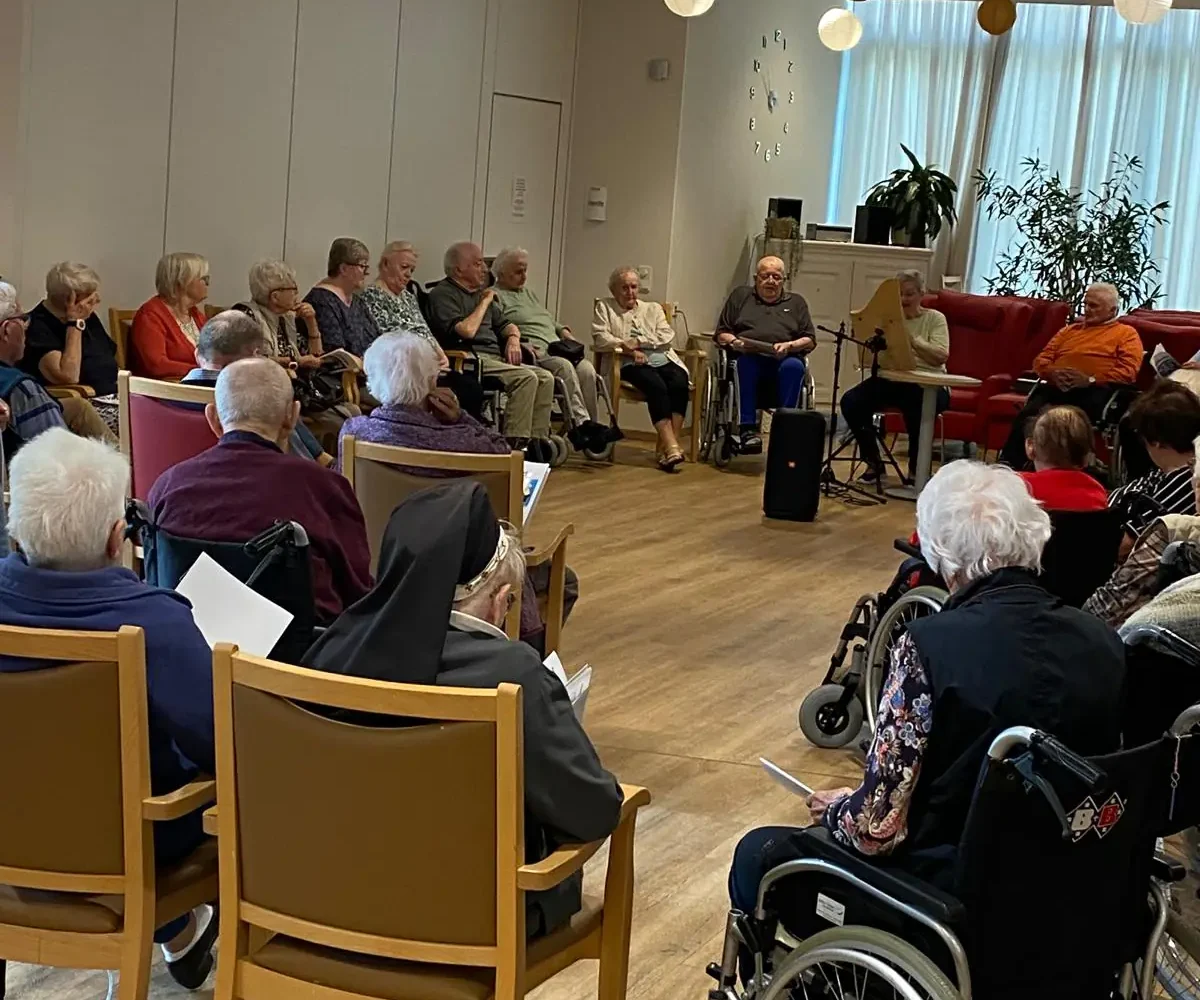 Ältere Menschen sitzen mit Liedblättern im Kreis, während eine Frau ein Saiteninstrument spielt.