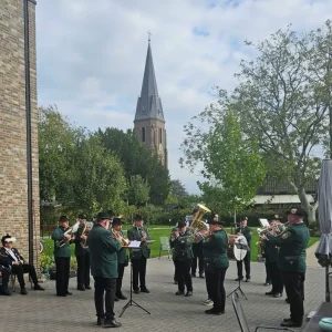 Eine Blaskapelle spielt vor einem Backsteingebäude. Im Hintergrund ragt ein Kirchturm über die Bäume.