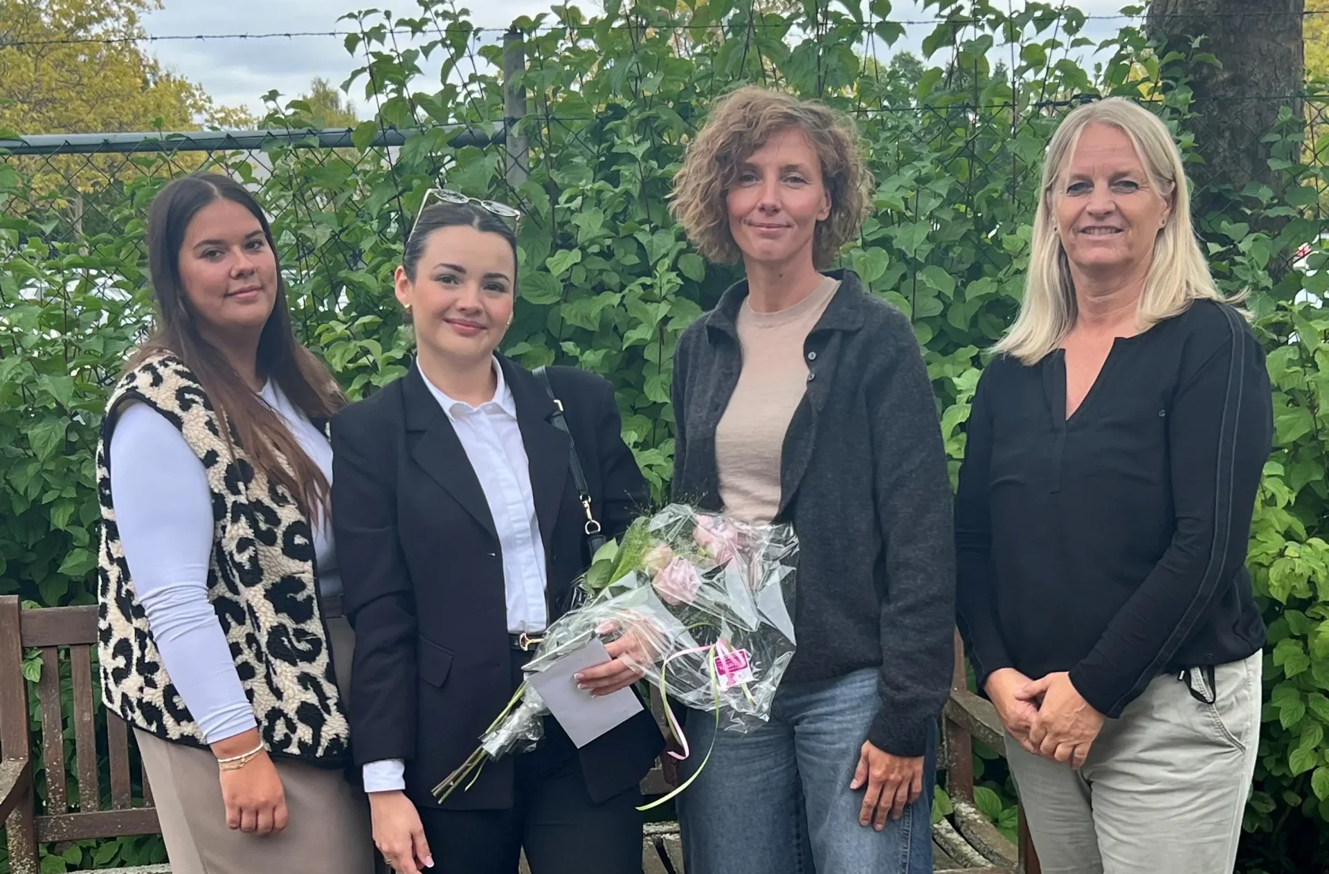Vier Frauen stehen vor einer Holzbank im Grünen. Die zweite Frau von links trägt einen schwarzen Anzug und hält einen Blumenstrauß.