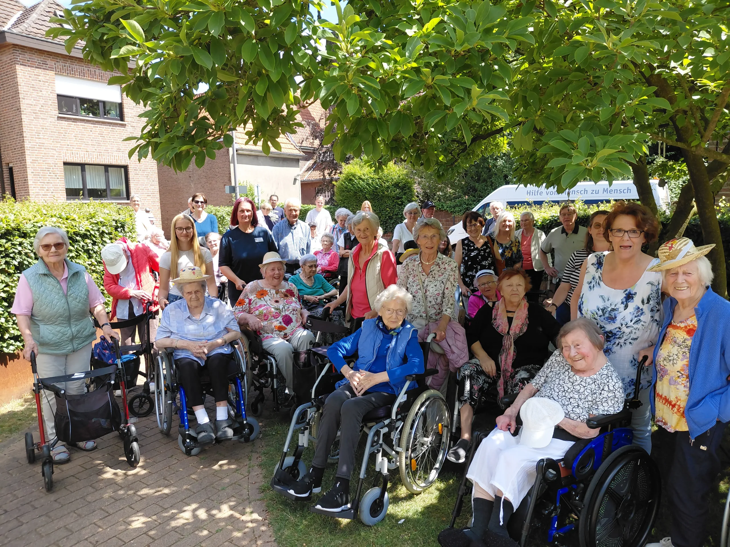 Eine große Gruppe älterer Menschen und Begleitpersonen posiert unter einem Baum, einige mit Rollstühlen und Rollatoren.