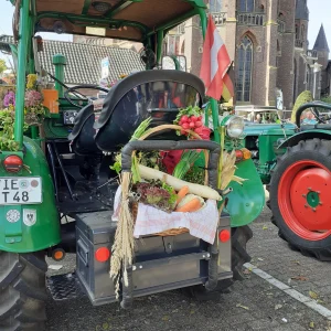 Ein grüner Oldtimertraktor steht vor einer Kirche, geschmückt mit einem Korb voller Gemüse und Getreideähren.