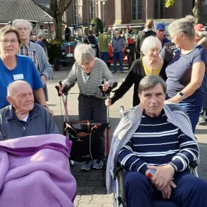 Ältere Menschen mit Rollstühlen und Rollator besuchen mit Begleitpersonen eine Traktorenausstellung vor einer Kirche.