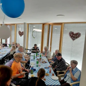 Ältere Menschen sitzen an einer langen Tafel mit blau-weißer Tischdecke, Getränken und Snacks unter Luftballons.