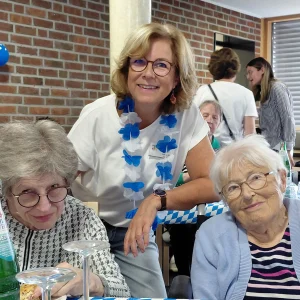 Drei Frauen lächeln an einem gedeckten Tisch in die Kamera. Die Frau in der Mitte trägt eine blau-weiße Blumenkette.