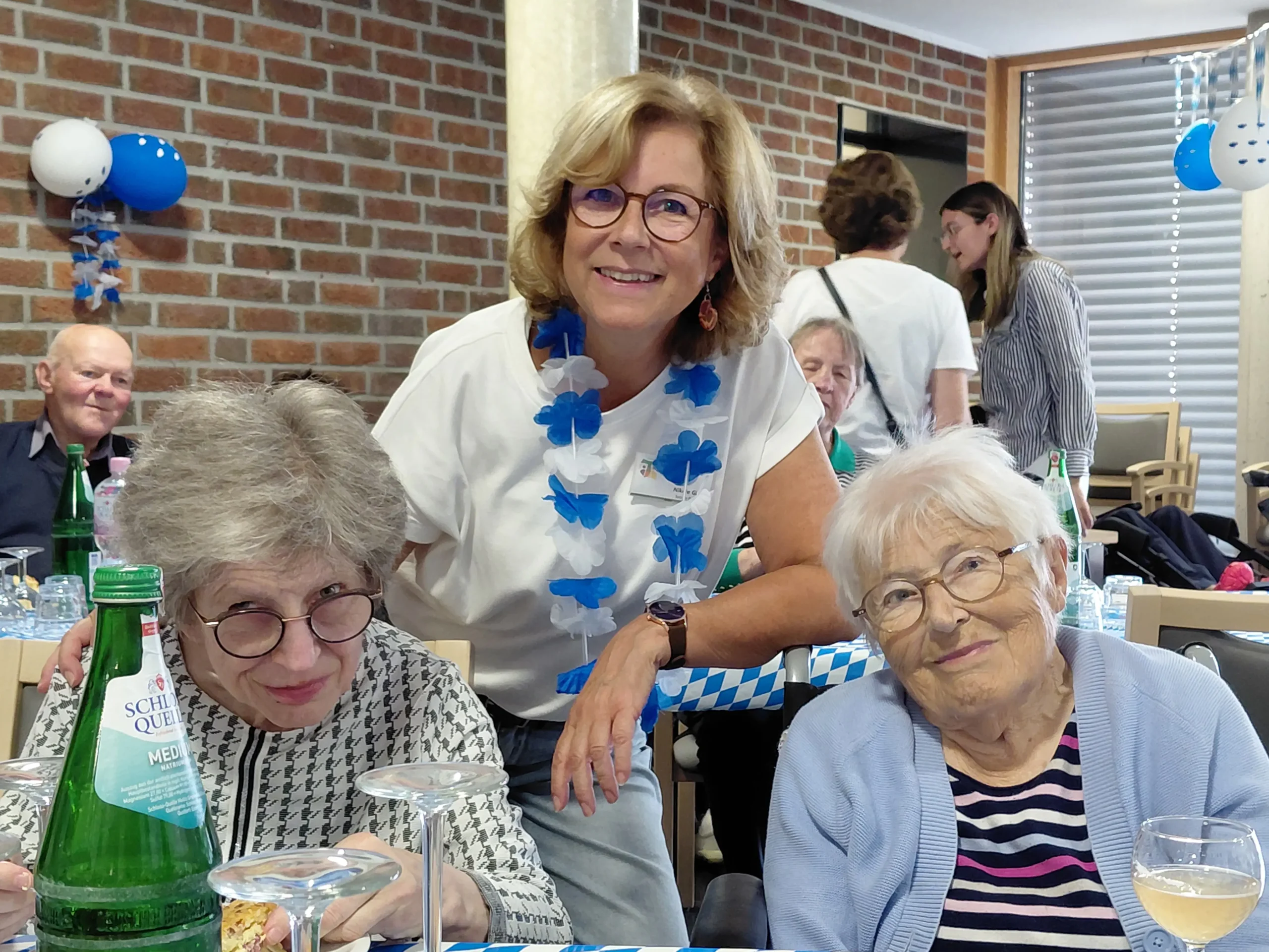 Drei Frauen lächeln an einem gedeckten Tisch in die Kamera. Die Frau in der Mitte trägt eine blau-weiße Blumenkette.