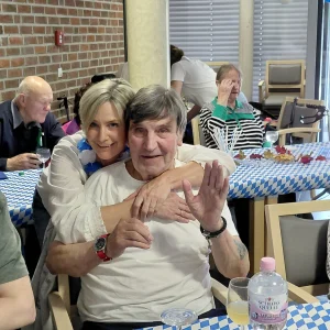 Eine Frau umarmt einen sitzenden älteren Mann, der in die Kamera winkt. Beide lächeln.