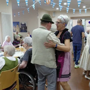 Ein Mann mit grünem Hut und eine Frau im Dirndl tanzen zwischen den Tischen unter blau-weißen Wimpeln.