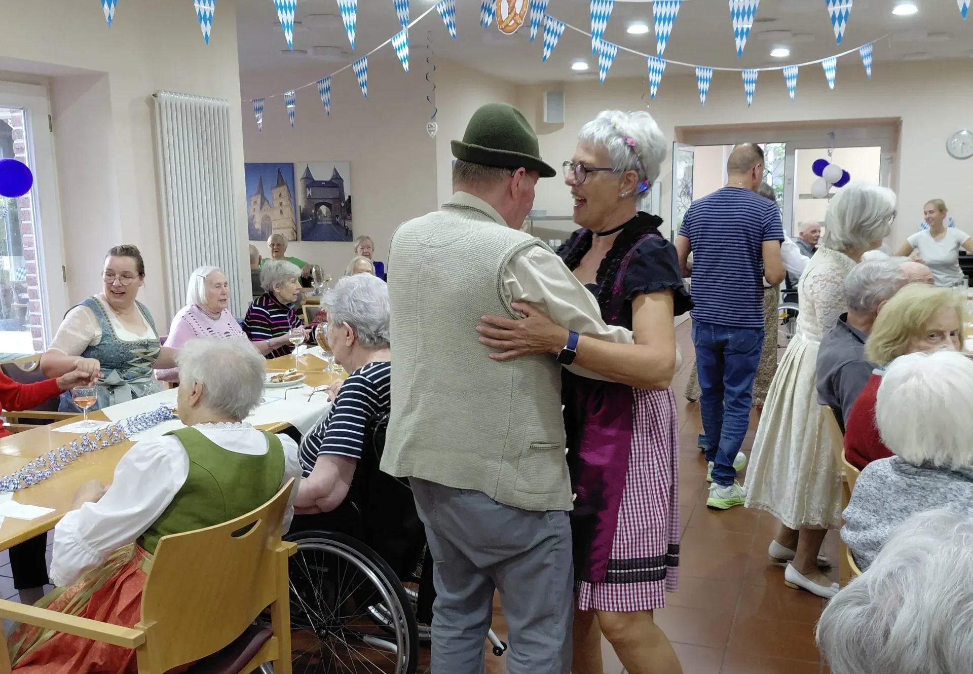 Ein Mann mit grünem Hut und eine Frau im Dirndl tanzen zwischen den Tischen unter blau-weißen Wimpeln.