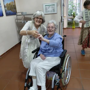 Eine Frau im weißen Dirndl und eine ältere Frau im Rollstuhl halten sich lächelnd an der Hand. Dahinter spielt Akkordeonmusik.