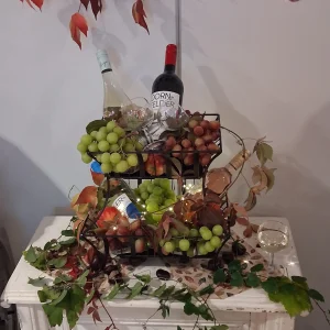 Weinflaschen, Gläser und Trauben stehen in einem Metallkorb, geschmückt mit Herbstlaub, Efeu und einer Lichterkette.