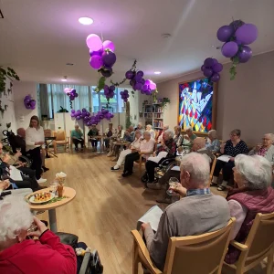 Ältere Menschen sitzen mit Gläsern und Liedblättern im Kreis. Lila Luftballons in Traubenform schmücken den Raum.