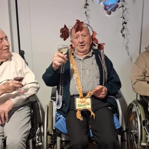 Drei ältere Männer sitzen in Rollstühlen. Der Mann in der Mitte trägt einen Blätterkranz und hebt sein Weinglas.