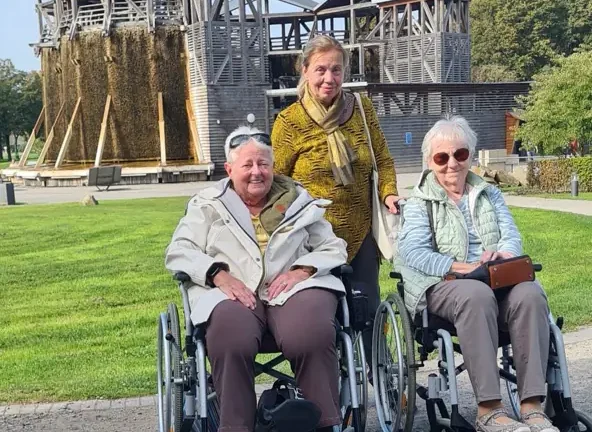 Zwei ältere Frauen im Rollstuhl und eine stehende Frau posieren vor einem Gradierwerk im Grünen.