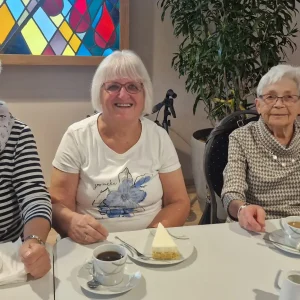 Vier Frauen sitzen nebeneinander an einem Tisch mit Kaffeetassen und Tortenstücken und blicken in die Kamera.