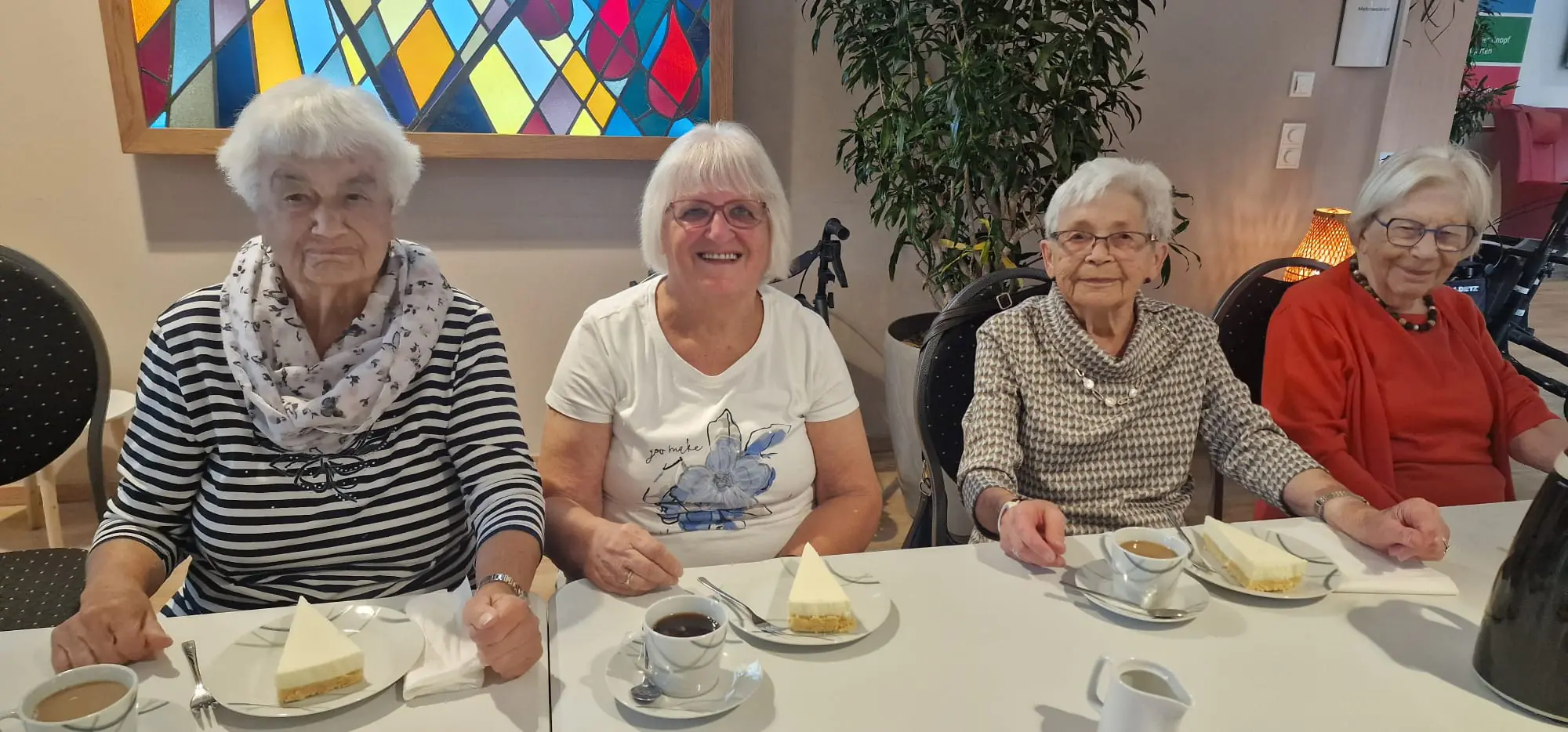 Vier Frauen sitzen nebeneinander an einem Tisch mit Kaffeetassen und Tortenstücken und blicken in die Kamera.