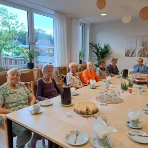 Sieben Frauen sitzen an einer gedeckten Kaffeetafel mit verschiedenen Torten vor einer großen Fensterfront.