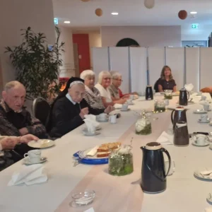 Eine Gruppe älterer Menschen sitzt an einer langen Tafel mit Kaffee, Kuchen und Blumen in dekorierten Gläsern.