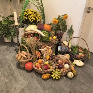 Körbe mit Obst und Gemüse, Kürbisse und Getreideähren sind neben Blumen und einer brennenden Kerze arrangiert.