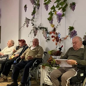 Vier ältere Männer sitzen vor einer Wand mit Traubendekoration. Einer liest ein Blatt, andere halten Gläser.