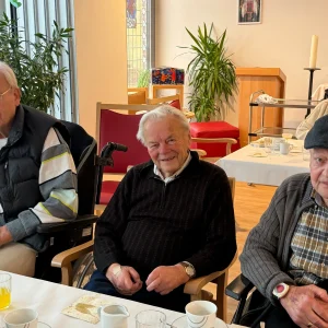 Drei ältere Männer sitzen gemeinsam an einer gedeckten Tafel im Gemeinschaftsraum eines Seniorenheims.